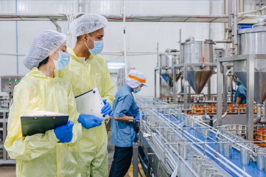 Hai chuyên viên kiểm tra dây chuyền sản xuất thực phẩm tại nhà máy, minh họa cho việc phân loại giá thành sản phẩm theo thời điểm tính toán.